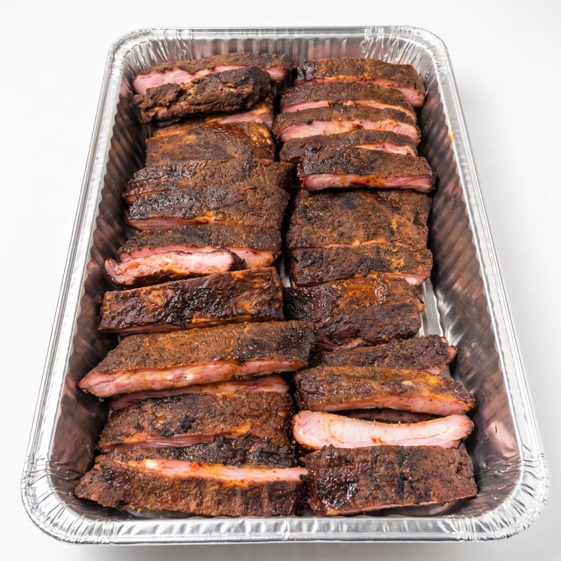 Close-up of sliced, smoked ribs with a dark crust and pink meat in a foil catering pan.