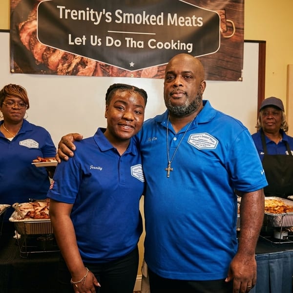 Trenity's Smoked Meats staff, including a man and woman in blue shirts, stand by buffet food.