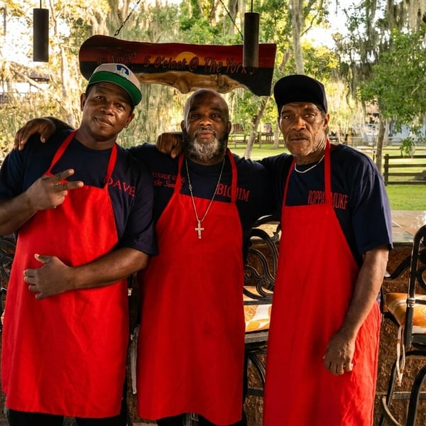 Three men in red aprons and dark shirts stand together at an outdoor bar by a lake.