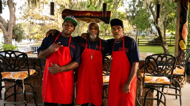 Three men in red aprons and dark shirts stand together at an outdoor bar by a lake.